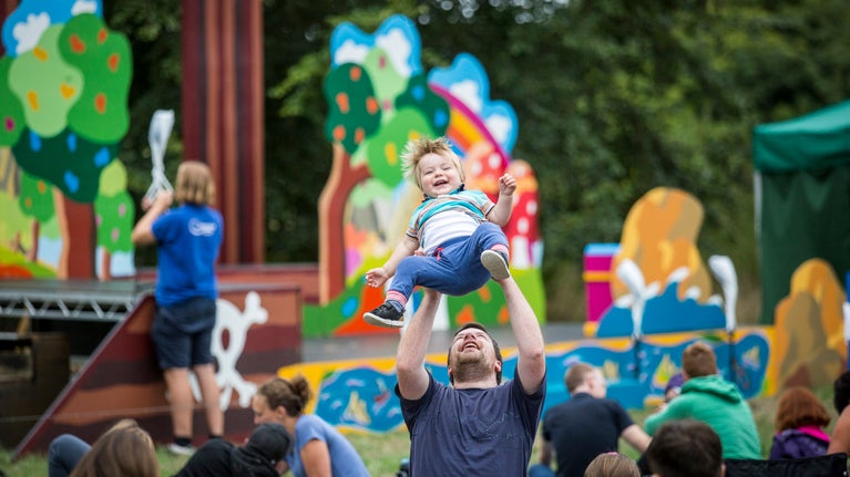 A child laughing while being held in the air by a adult at an open air theatre performance